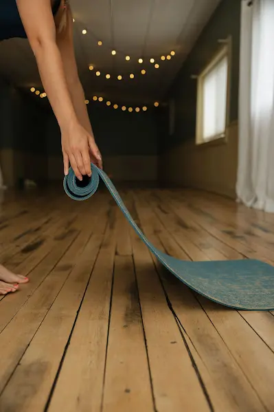 Person rolling up a blue yoga mat on wooden floor with string lights.