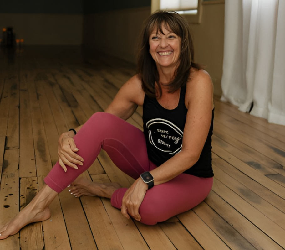 A smiling woman in pink leggings sitting on a wooden floor.